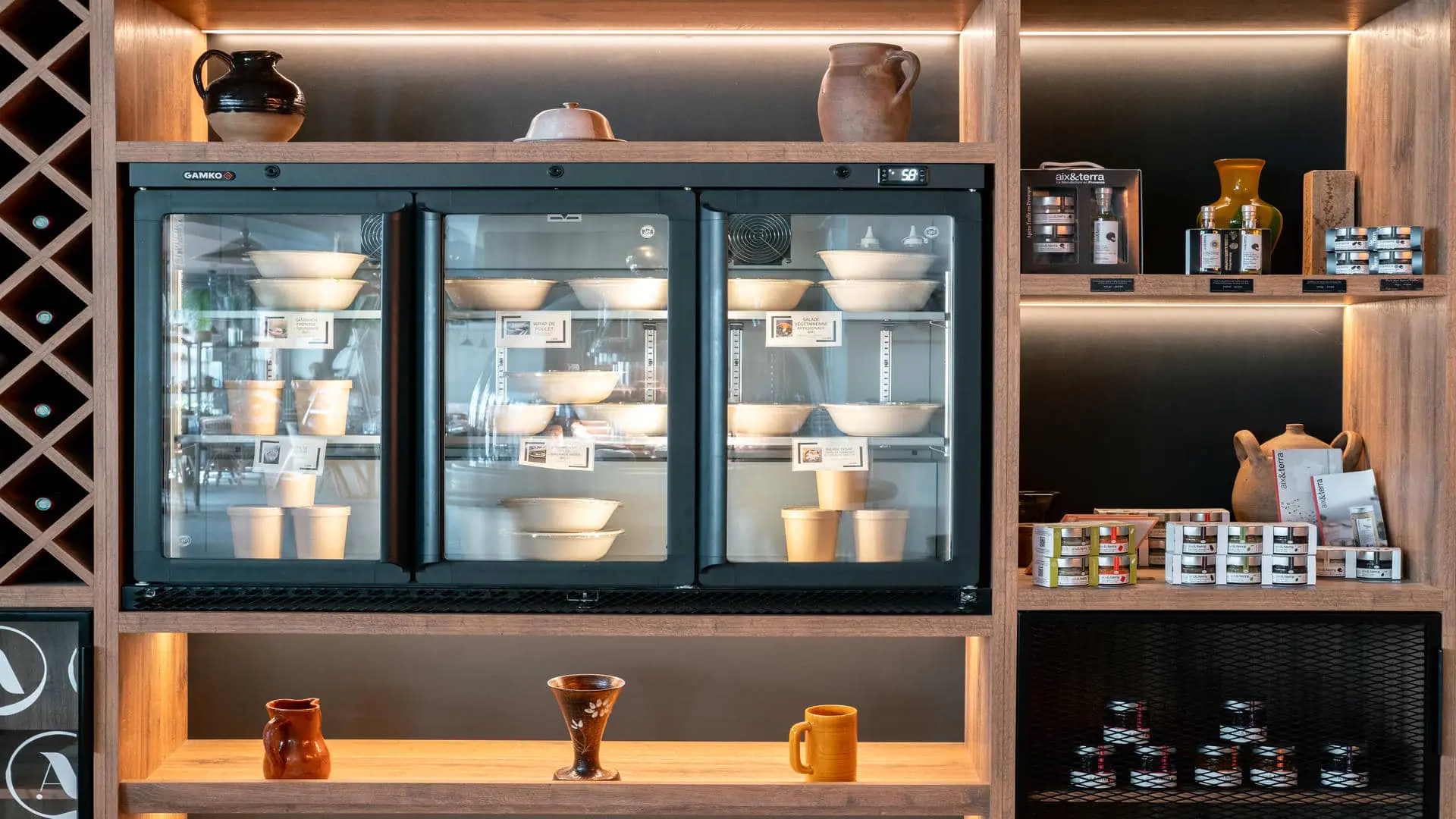 en::Gourmet shop with wooden, wire and open shelves displaying wine bottles, tableware décor, gift boxes, refrigerated takeaway dishes, jams and artisan condiments. fr::Boutique épicerie avec étagères en bois, grillagées et ouvertes, bouteilles de vin, arts de la table, coffrets cadeaux, plats à emporter réfrigérés, confitures et condiments artisanaux.