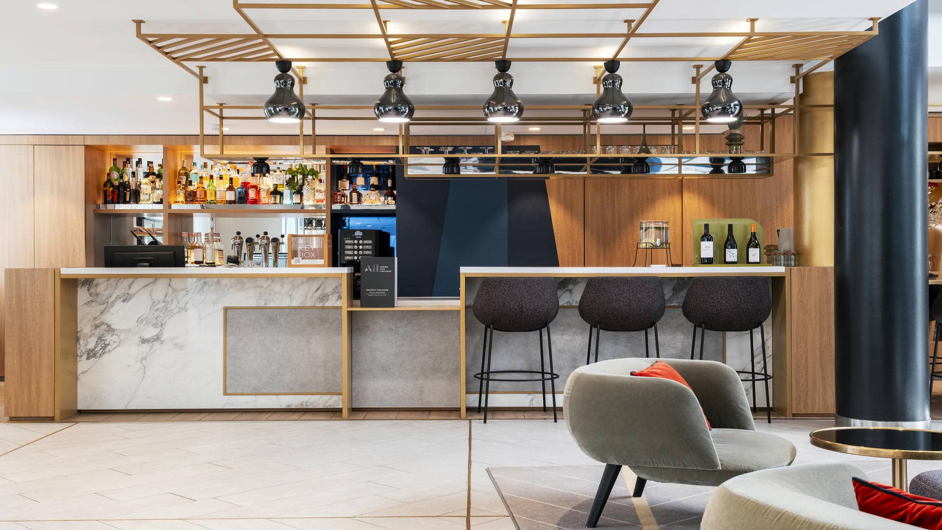 en:: Bar counter in the seminar hotel lobby, made of wood and grey-and-white marble, with black ceiling lights and a wooden backdrop. fr::Comptoir de bar dans le lobby de l’hôtel séminaire, en bois et marbre gris et blanc, avec luminaires noirs au plafond et arrière-plan en bois.