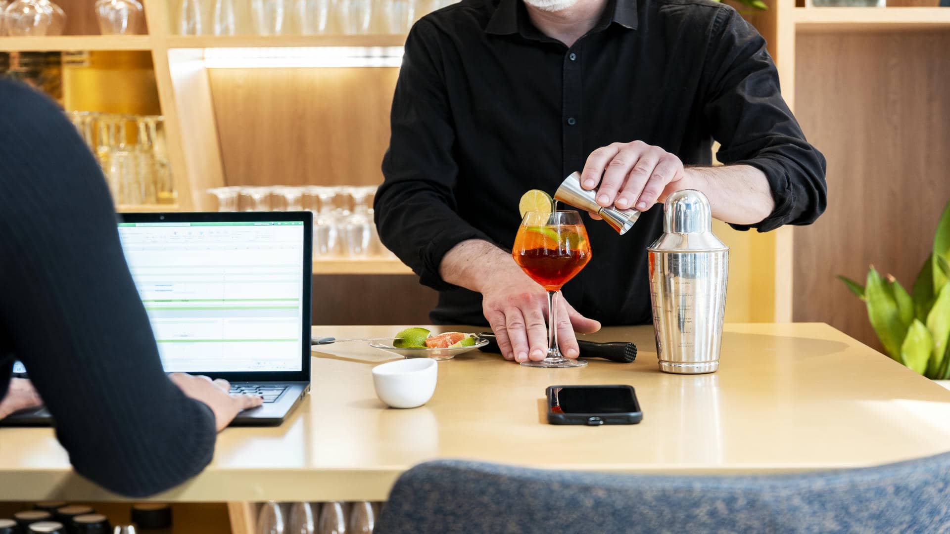 en::Bartender preparing a colourful cocktail with a shaker and jigger, facing a guest with an open laptop at the wooden counter of the seminar restaurant. fr::Barman préparant un cocktail coloré au shaker face à un client avec ordinateur portable ouvert au comptoir en bois du restaurant séminaire.