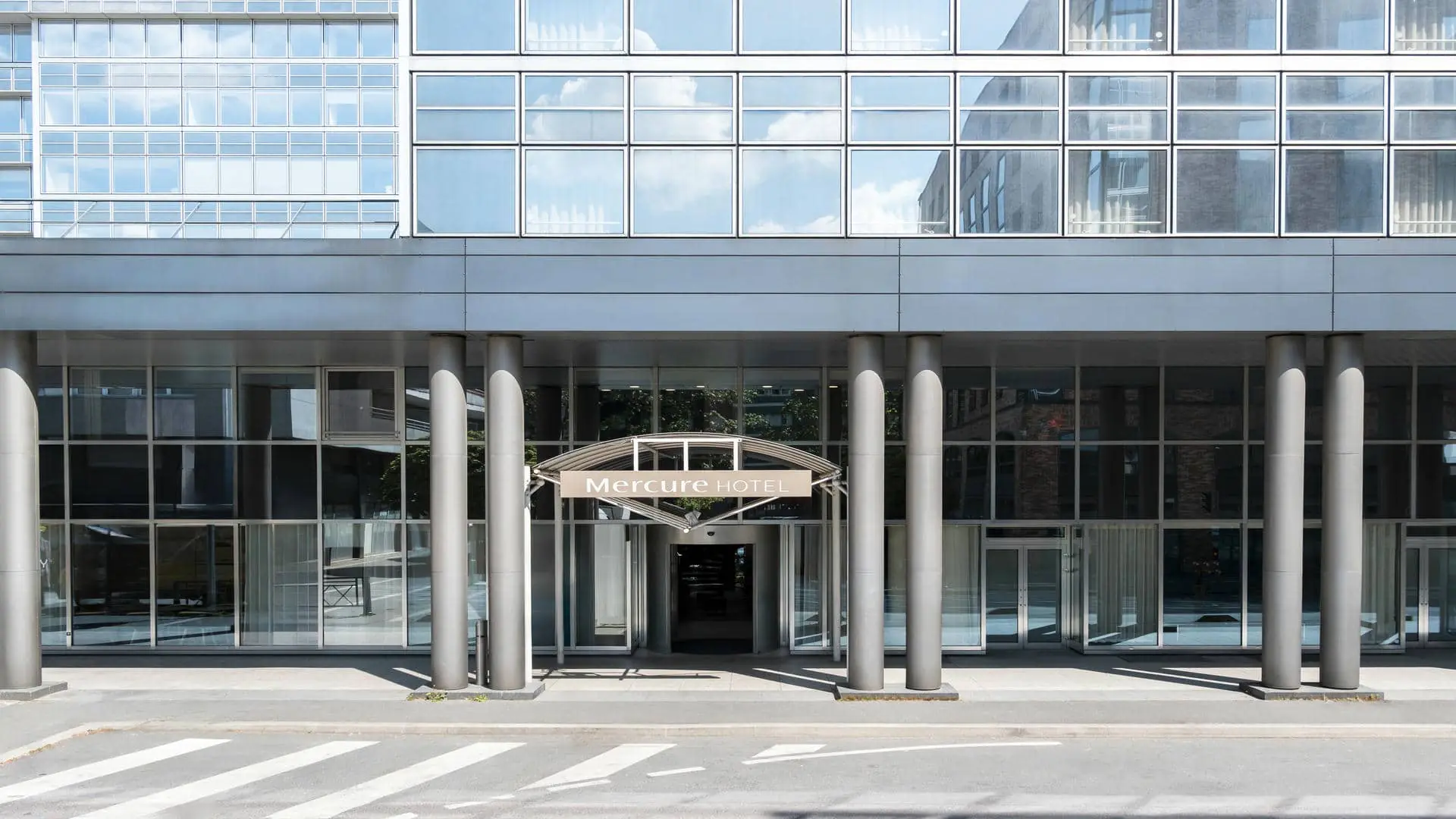 en::Glass façade of the hotel with the Mercure Hotel sign at the entrance and a visible revolving door. fr::Façade vitrée de l’hôtel avec enseigne Mercure Hotel en devanture et porte tournante visible.