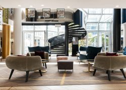 en::Seminar hotel lobby with a spiral staircase to the upper floors, view of the inner courtyard, bar on the left and Mercure branded wall on the right. fr::Lobby de l’hôtel séminaire avec escalier en spirale vers les étages, vue sur la cour intérieure, bar à gauche et mur de marque Mercure à droite.