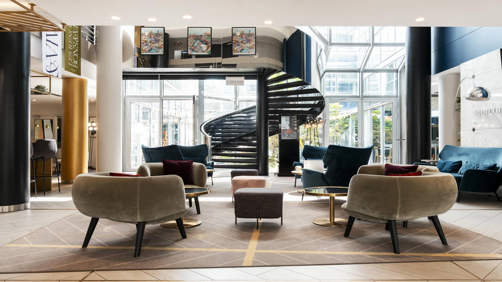 en::Seminar hotel lobby with a spiral staircase to the upper floors, view of the inner courtyard, bar on the left and Mercure branded wall on the right. fr::Lobby de l’hôtel séminaire avec escalier en spirale vers les étages, vue sur la cour intérieure, bar à gauche et mur de marque Mercure à droite.