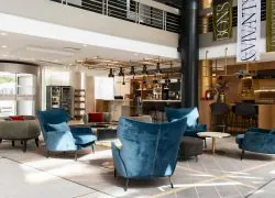 en::Seminar hotel lobby with a visible mezzanine, bar on the right, and central lounge with varied seating and gold coffee tables. fr::Lobby de l’hôtel séminaire avec mezzanine visible, bar à droite et salon central avec assises variées et tables basses dorées.