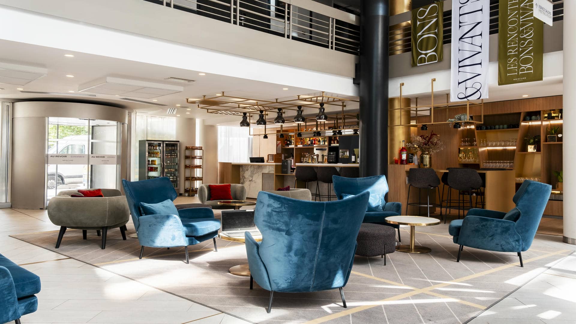 en::Seminar hotel lobby with a visible mezzanine, bar on the right, and central lounge with varied seating and gold coffee tables. fr::Lobby de l’hôtel séminaire avec mezzanine visible, bar à droite et salon central avec assises variées et tables basses dorées.