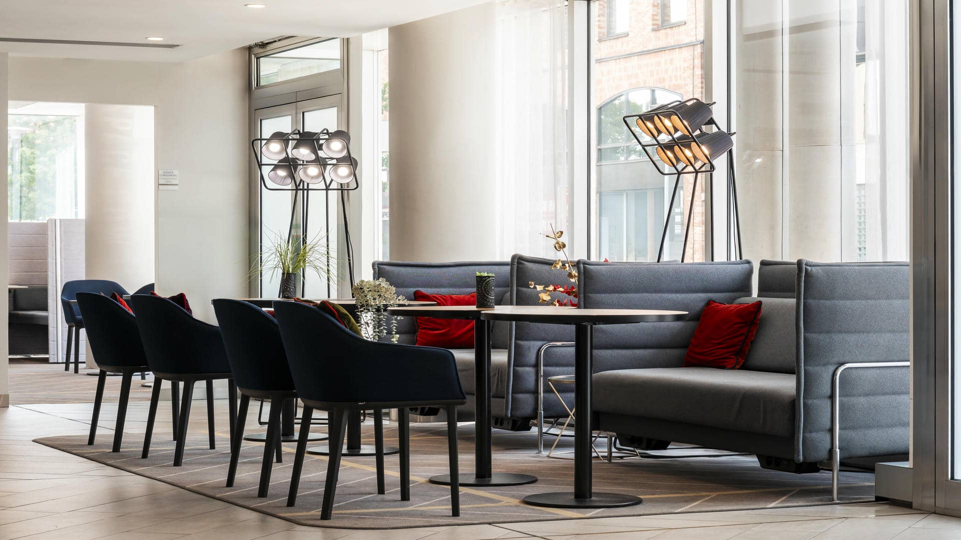 en::Coworking space with two large grey fabric armchairs, each facing a round work table and a blue velvet chair, plus a more private coworking area with partitions in the background. fr::Espace coworking avec deux grands fauteuils gris en tissu, chacun face à une table de travail ronde et un siège bleu en velours, avec un espace plus intimiste cloisonné en arrière-plan.