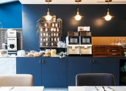 en::Seminar hotel breakfast room viewed from the tables toward the open-kitchen buffet along a blue wall, with a DeLaFerme yogurt machine, coffee machine and jam shelves. fr::Salle petit déjeuner de l’hôtel séminaire, vue depuis les tables vers le buffet façon cuisine ouverte le long d’un mur bleu, avec machine à yaourt DeLaFerme, machine à café et étagères à confitures