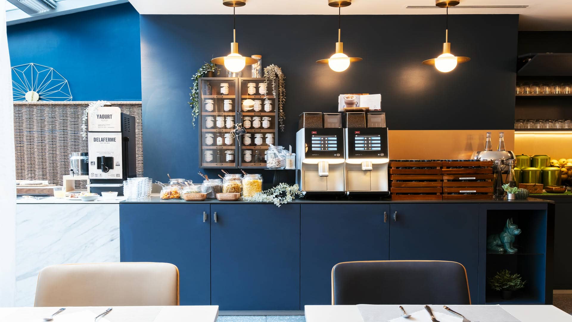en::Seminar hotel breakfast room viewed from the tables toward the open-kitchen buffet along a blue wall, with a DeLaFerme yogurt machine, coffee machine and jam shelves. fr::Salle petit déjeuner de l’hôtel séminaire, vue depuis les tables vers le buffet façon cuisine ouverte le long d’un mur bleu, avec machine à yaourt DeLaFerme, machine à café et étagères à confitures