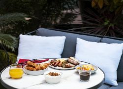 en::Breakfast on a terrace with grilled sausage, bacon, eggs, pastries, farm eco-yogurt and fruit, served on a round grey marble table with sofa seating, blue cushions and greenery in the background. fr::Petit déjeuner en terrasse avec saucisse grillée, bacon, œufs, viennoiseries, yaourt fermier écologique et fruits, servi sur table ronde en marbre gris avec canapé, coussins bleus et végétation en arrière-plan.