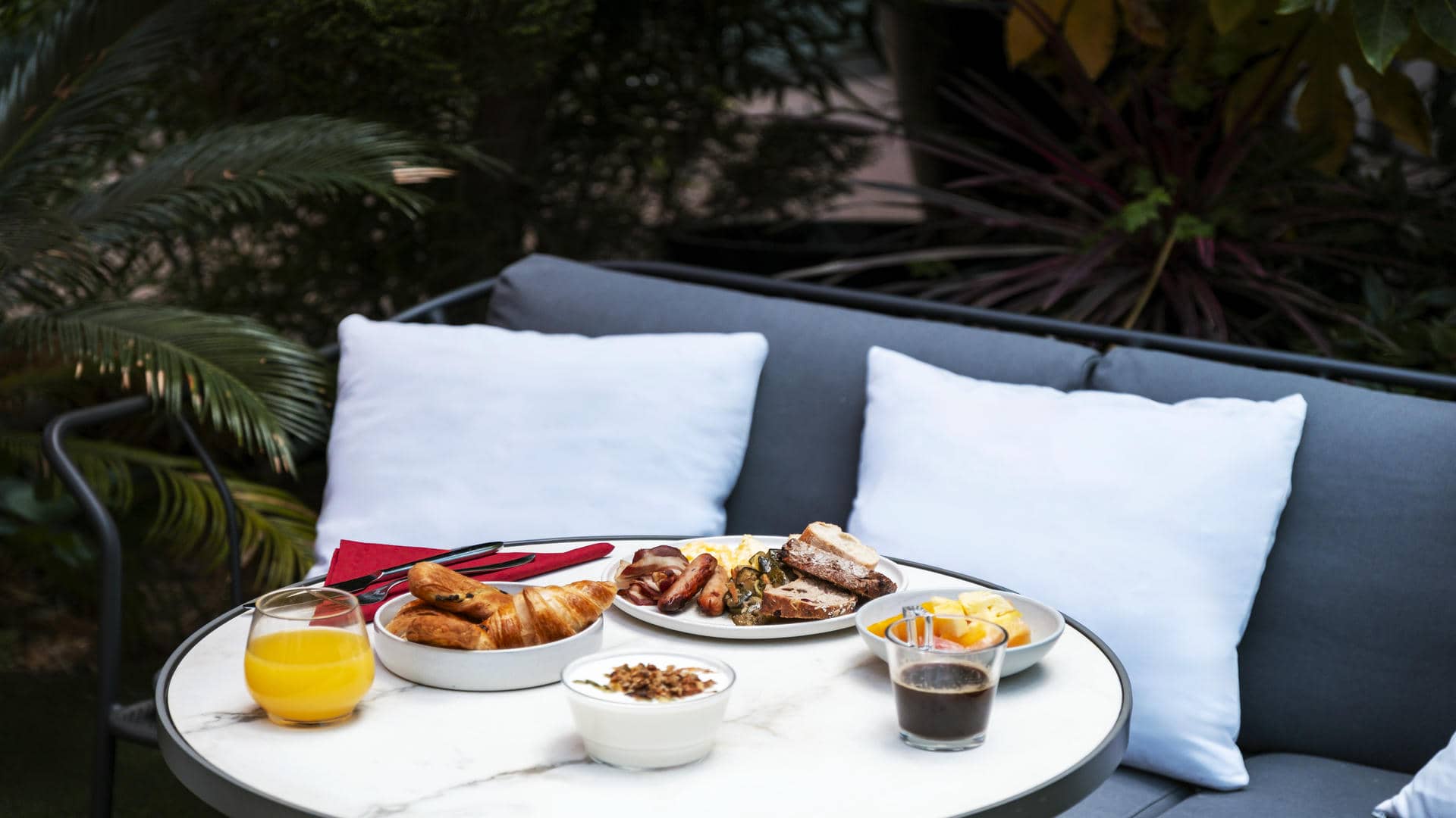 en::Breakfast on a terrace with grilled sausage, bacon, eggs, pastries, farm eco-yogurt and fruit, served on a round grey marble table with sofa seating, blue cushions and greenery in the background. fr::Petit déjeuner en terrasse avec saucisse grillée, bacon, œufs, viennoiseries, yaourt fermier écologique et fruits, servi sur table ronde en marbre gris avec canapé, coussins bleus et végétation en arrière-plan.
