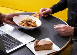 en::A person serves a gourmet plate to a seminar restaurant guest seated on a yellow sofa at a round black table with an open laptop. fr::Une personne sert une assiette gourmande à un client du restaurant séminaire, assis sur un canapé jaune devant une table ronde noire avec son ordinateur portable ouvert.