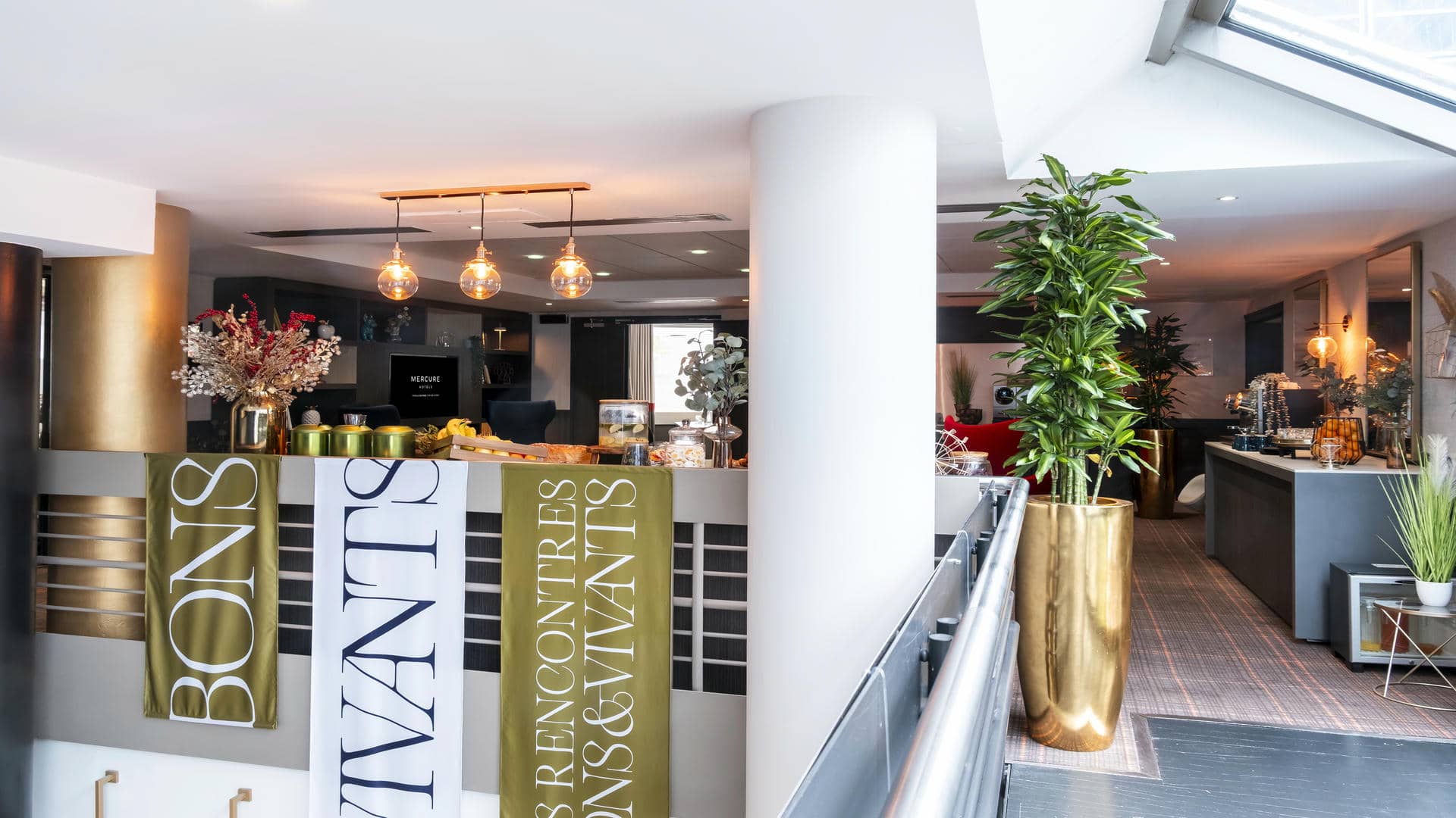 en::Lounge area above the staircase with a seminar break set up, and large banners hanging along the stairwell wall reading “Bons et Vivants.” fr::Espace salon au-dessus de l’escalier avec pause séminaire dressée et grandes banderoles “Bons et Vivants” le long du mur.