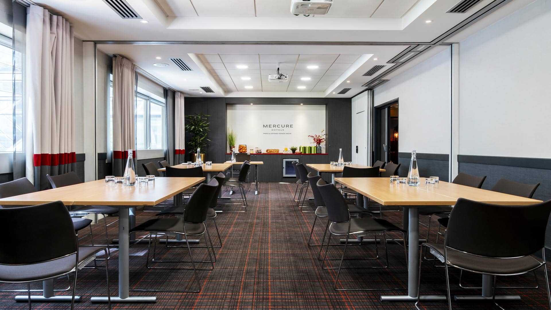 en::Bright, spotless meeting room with four table islands arranged on both sides of the room, projection screen at the front, seminar break below, and windows overlooking the city. fr::Salle de réunion lumineuse et impeccable avec quatre îlots de tables de part et d’autre de la pièce, écran de projection en face, pause séminaire en dessous et fenêtres avec vue sur la ville.