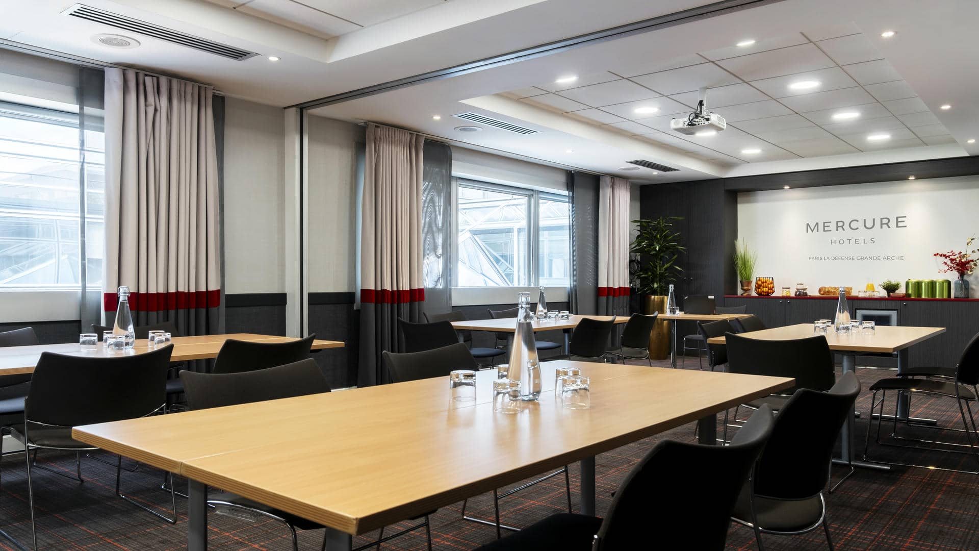 en::Bright, spotless meeting room with four table clusters arranged as islands, projection screen on the right wall, and windows overlooking the city buildings. fr::Salle de réunion lumineuse et impeccable avec quatre îlots de tables, écran de projection sur le mur droit et fenêtres avec vue sur la ville.