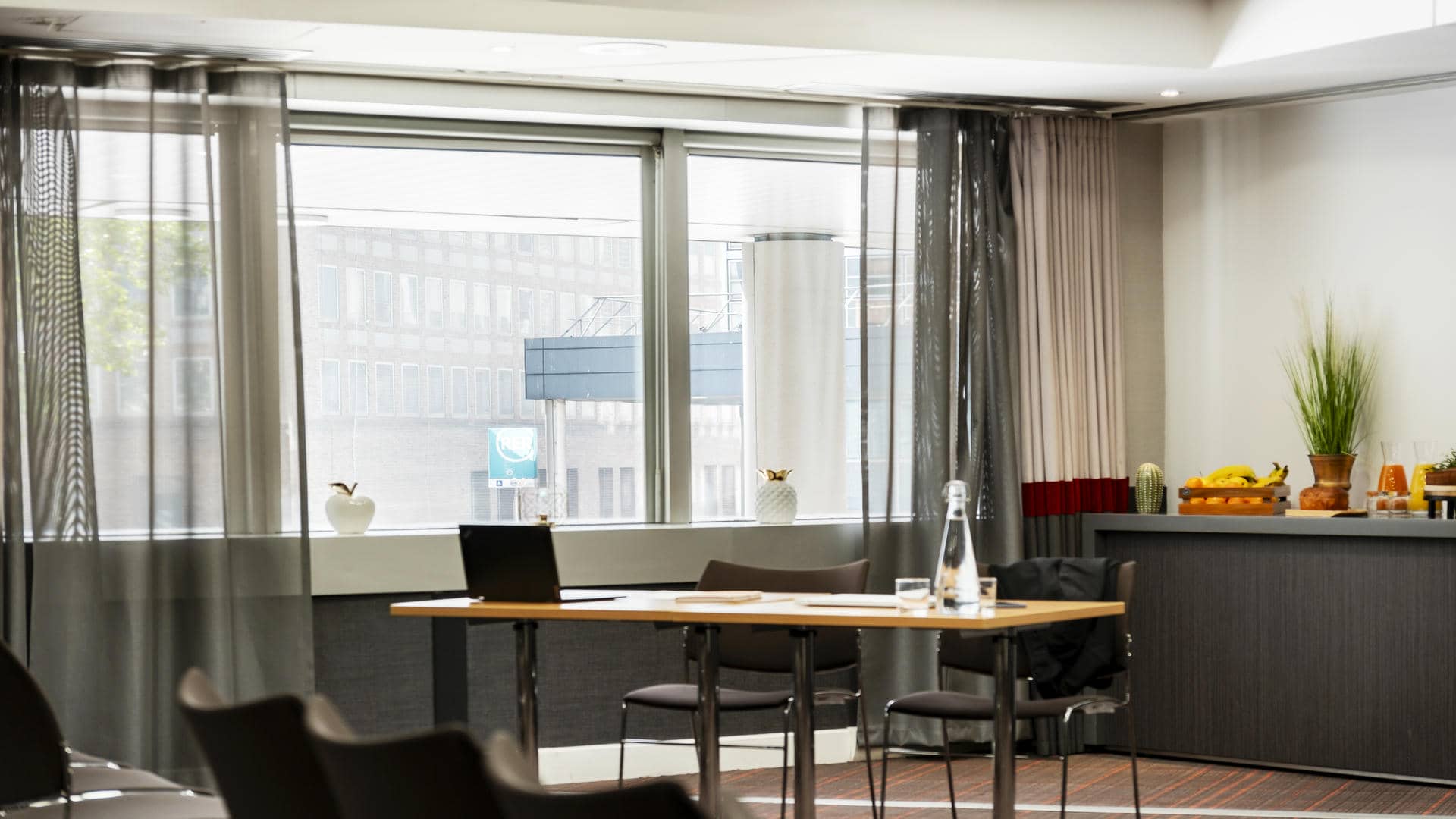 en::Meeting room with tables facing rows of theatre-style chairs, city and RER station views, with a seminar break set up in the background. fr::Salle de réunion avec tables face à des chaises en théâtre, vue sur la ville et la station RER, avec pause séminaire dressée en arrière-plan.