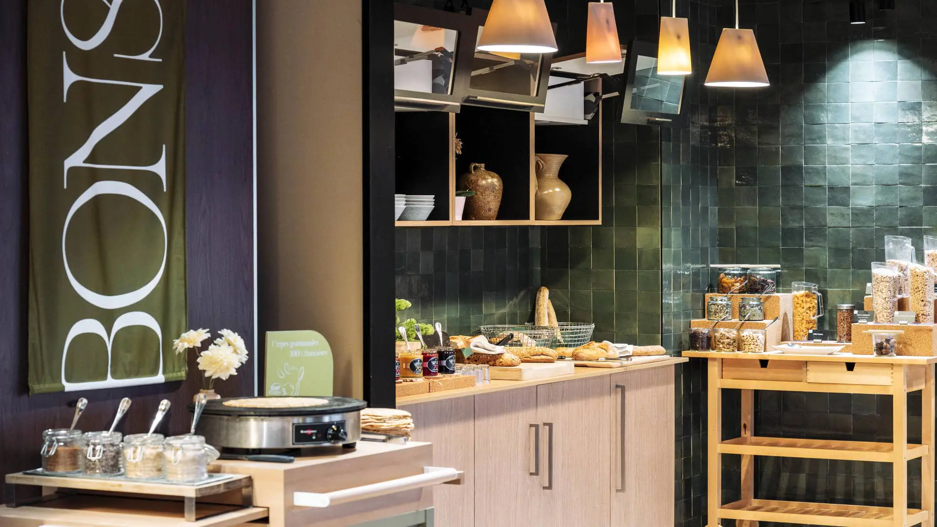 Buffet petit-déjeuner avec crêpière, toppings, confitures, céréales et produits en vrac, mur carrelé vert et meuble en bois.