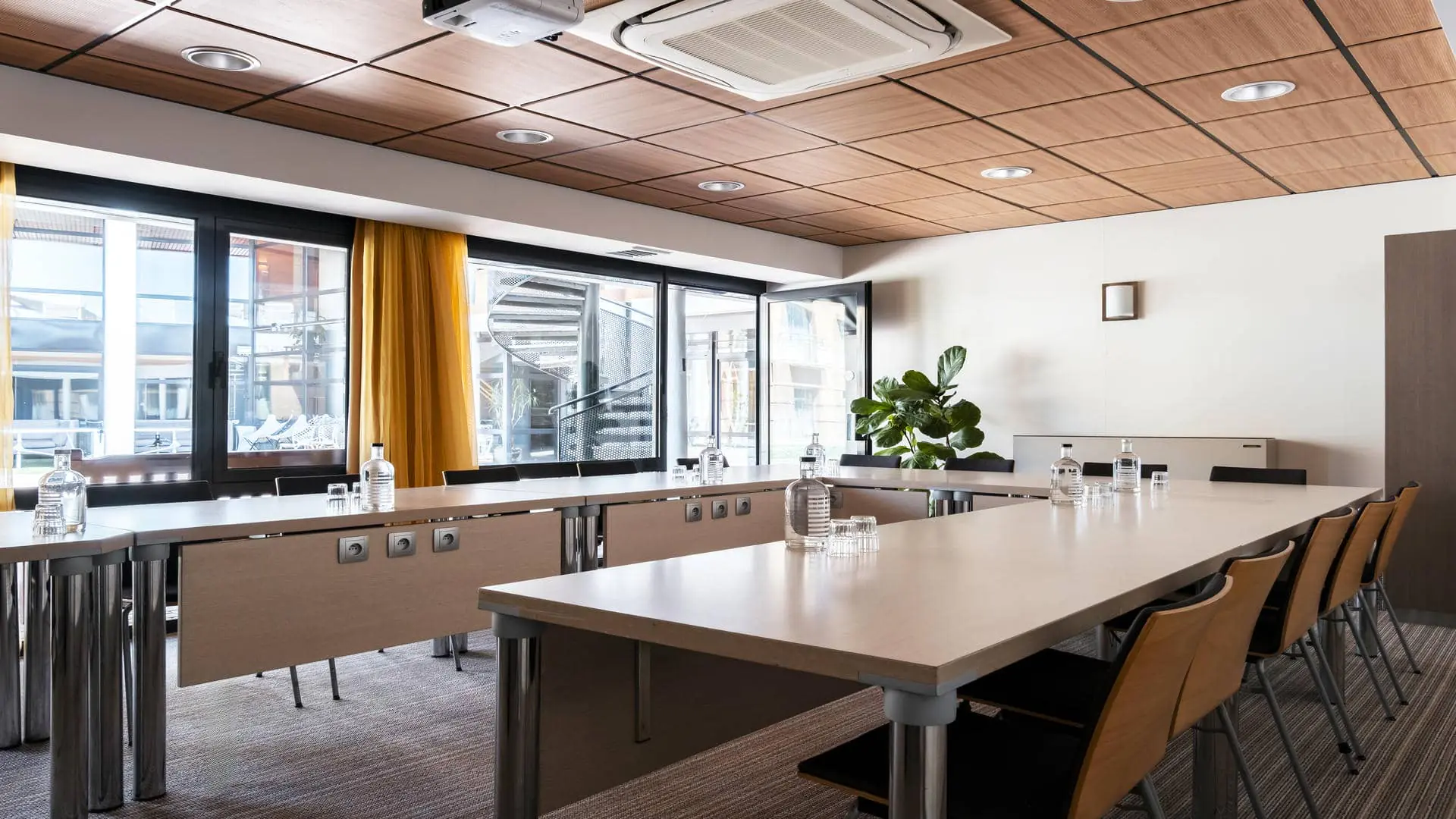 Salle de réunion avec tables en bois disposées en U, moquette beige et plafond en bois, voilages orangés et porte ouverte sur l’escalier en colimaçon et l’espace pause extérieur.