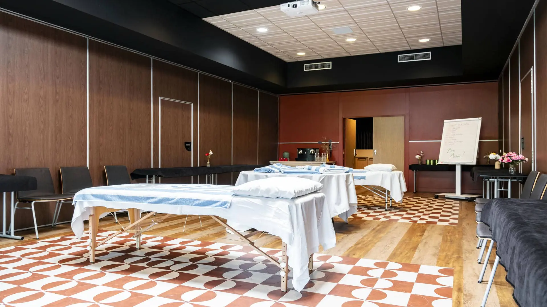 Salle de réunion avec sol en parquet et carrelage rouge et blanc, murs bordeaux, deux tables de massage disposées au centre et rangées de chaises de chaque côté de la salle.
