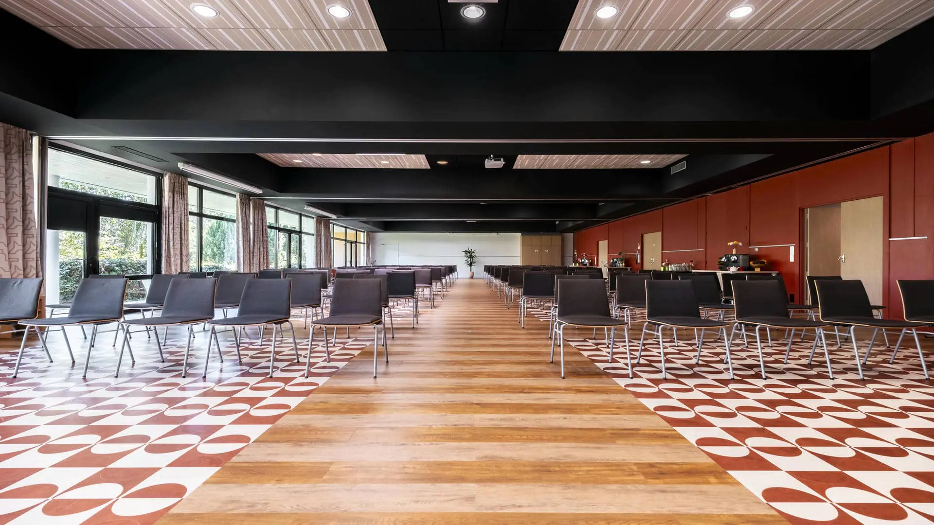 Salle de réunion avec sol en parquet et carrelage rouge et blanc, murs rouges et baies vitrées, rangées de chaises en bois de part et d’autre d’une allée, vue de face avec écran de projection au fond de la salle.
