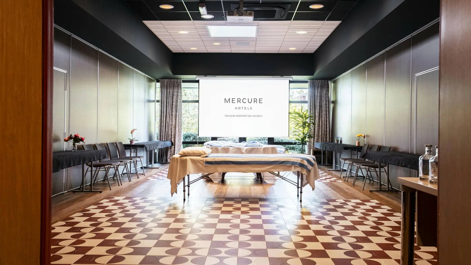Salle de réunion avec sol en parquet et carrelage rouge et blanc, murs bordeaux, deux tables de massage disposées au centre et rangées de chaises de chaque côté de la salle.