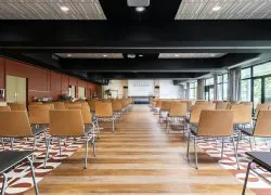 Salle de réunion avec sol en parquet et carrelage rouge et blanc, murs rouges à gauche et baies vitrées à droite, estrade en face et rangées de chaises en bois de part et d’autre d’une allée