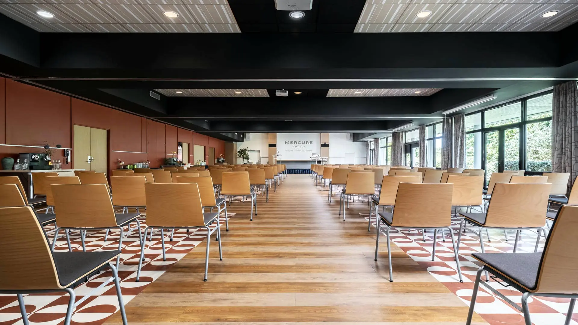 Salle de réunion avec sol en parquet et carrelage rouge et blanc, murs rouges à gauche et baies vitrées à droite, estrade en face et rangées de chaises en bois de part et d’autre d’une allée