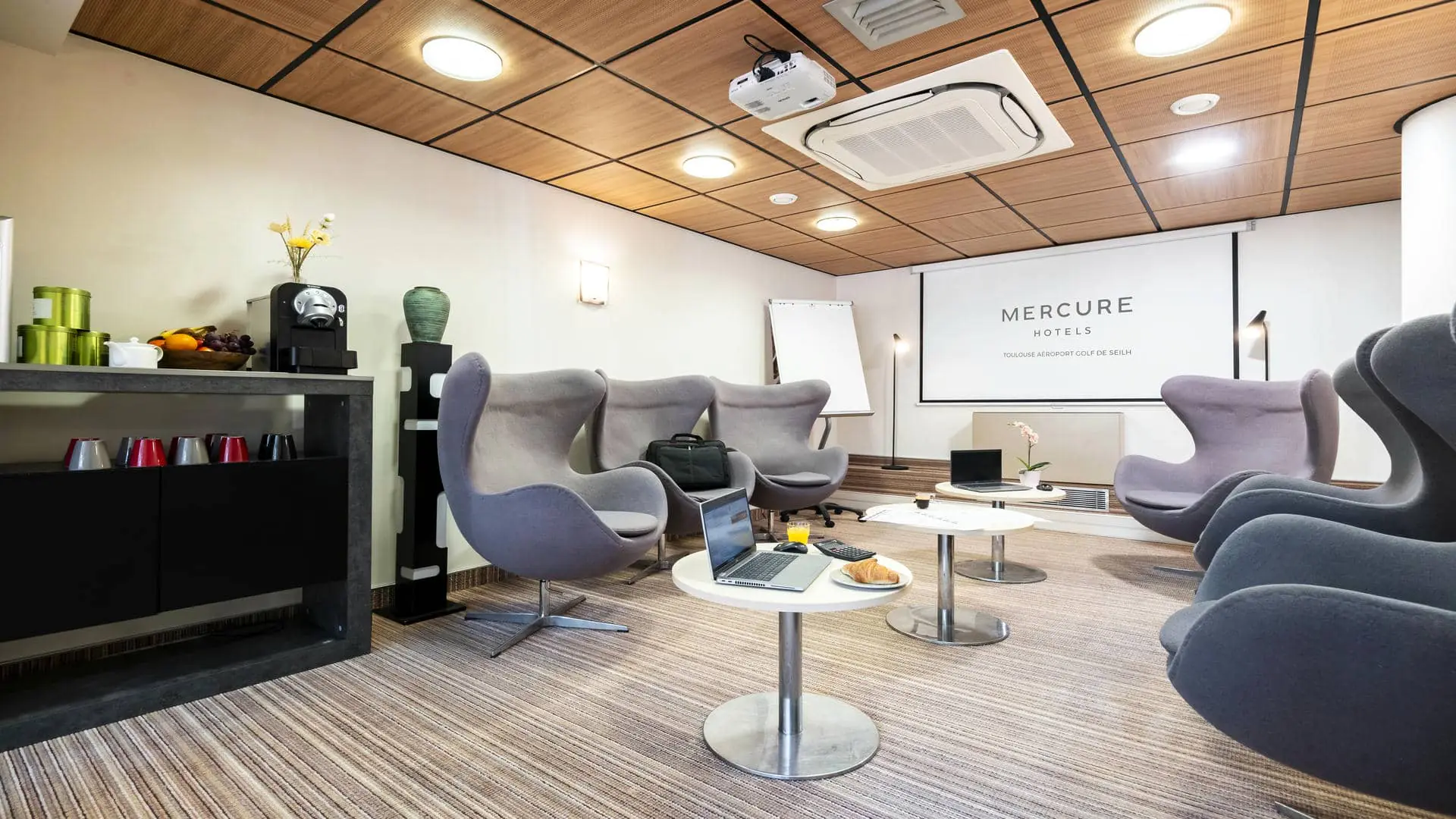 Larges fauteuils gris face à des tables basses, avec un écran de projection au fond de la salle et, sur l’un des côtés, un meuble aménagé pour une pause séminaire.