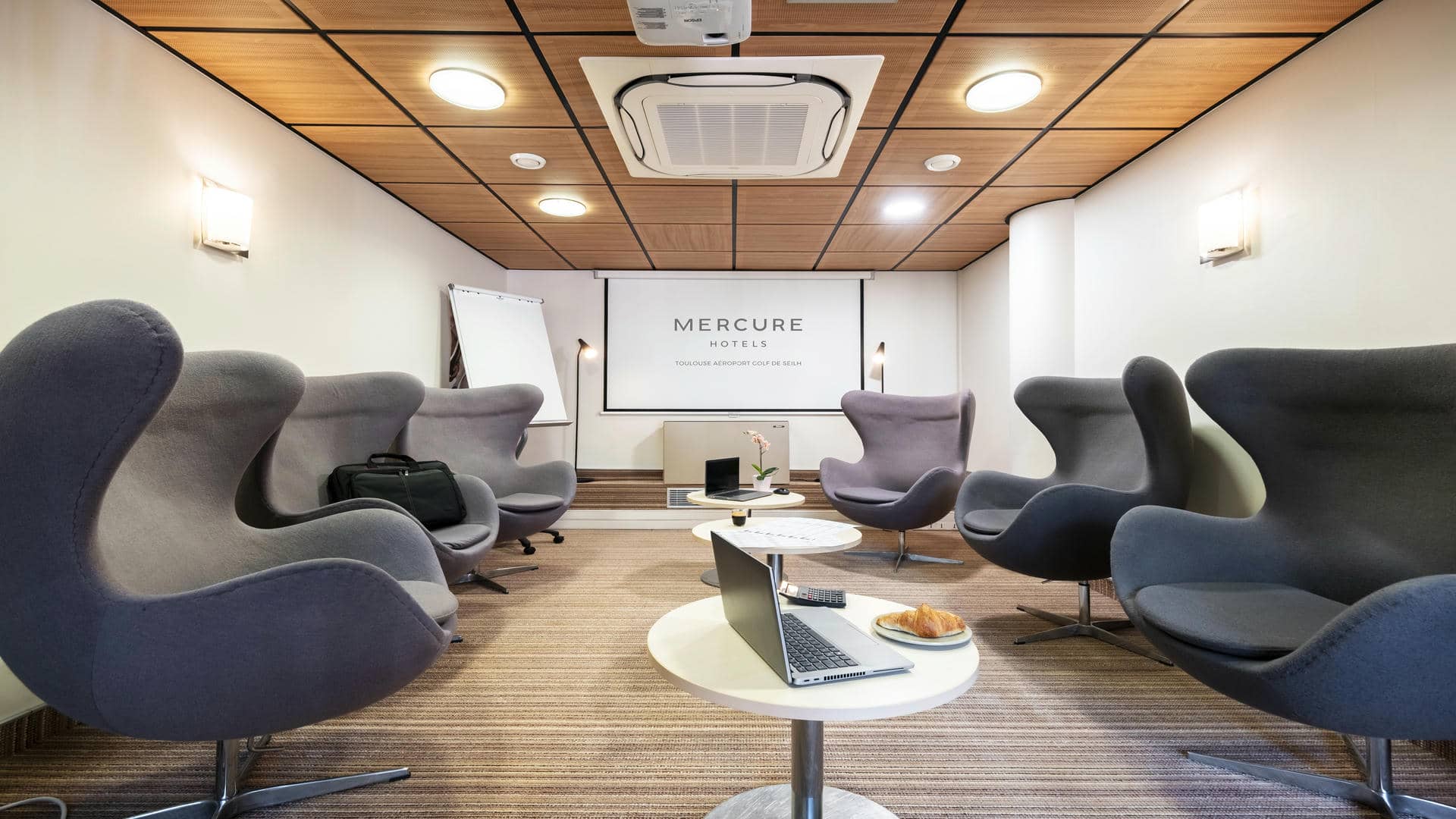 Larges fauteuils gris face à des tables basses, avec un écran de projection au fond de la salle et, sur l’un des côtés, un meuble aménagé pour une pause séminaire.