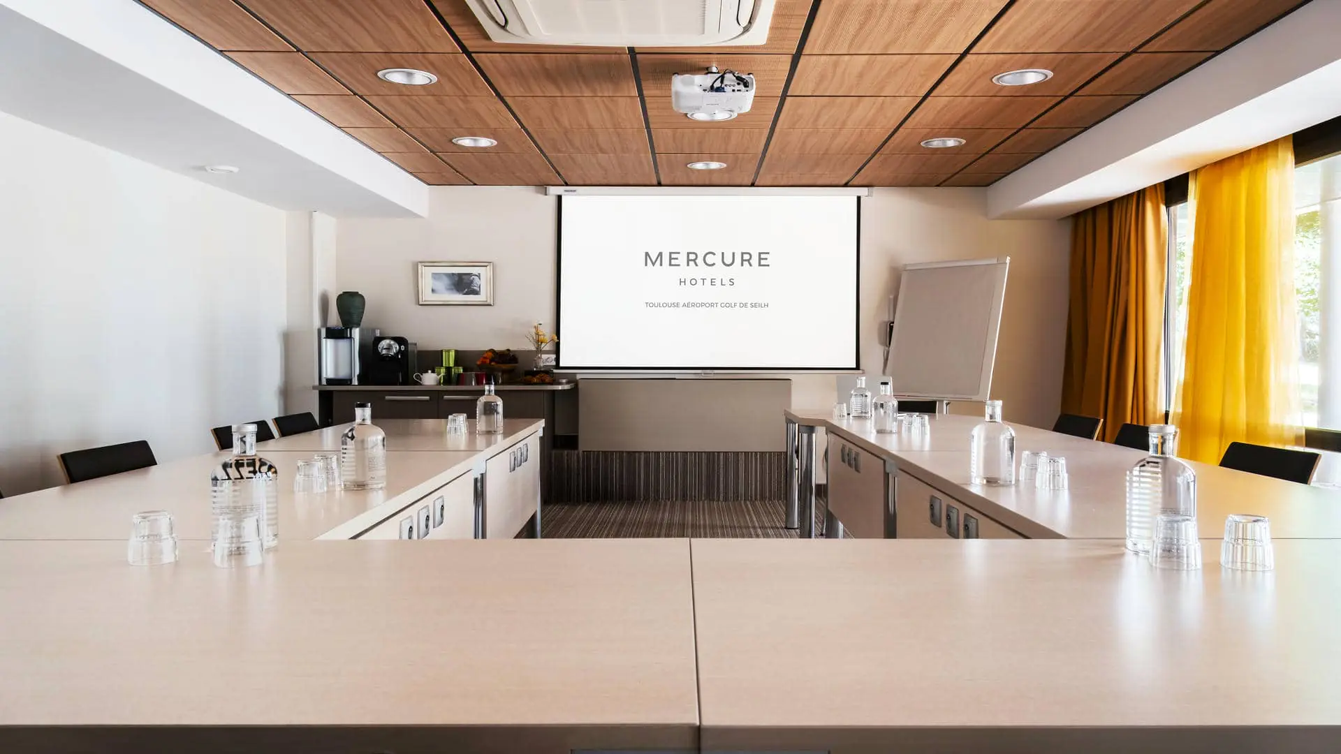 Salle de séminaire avec tables disposées en U et espace pause aménagé dans la salle sous l’écran de projection.