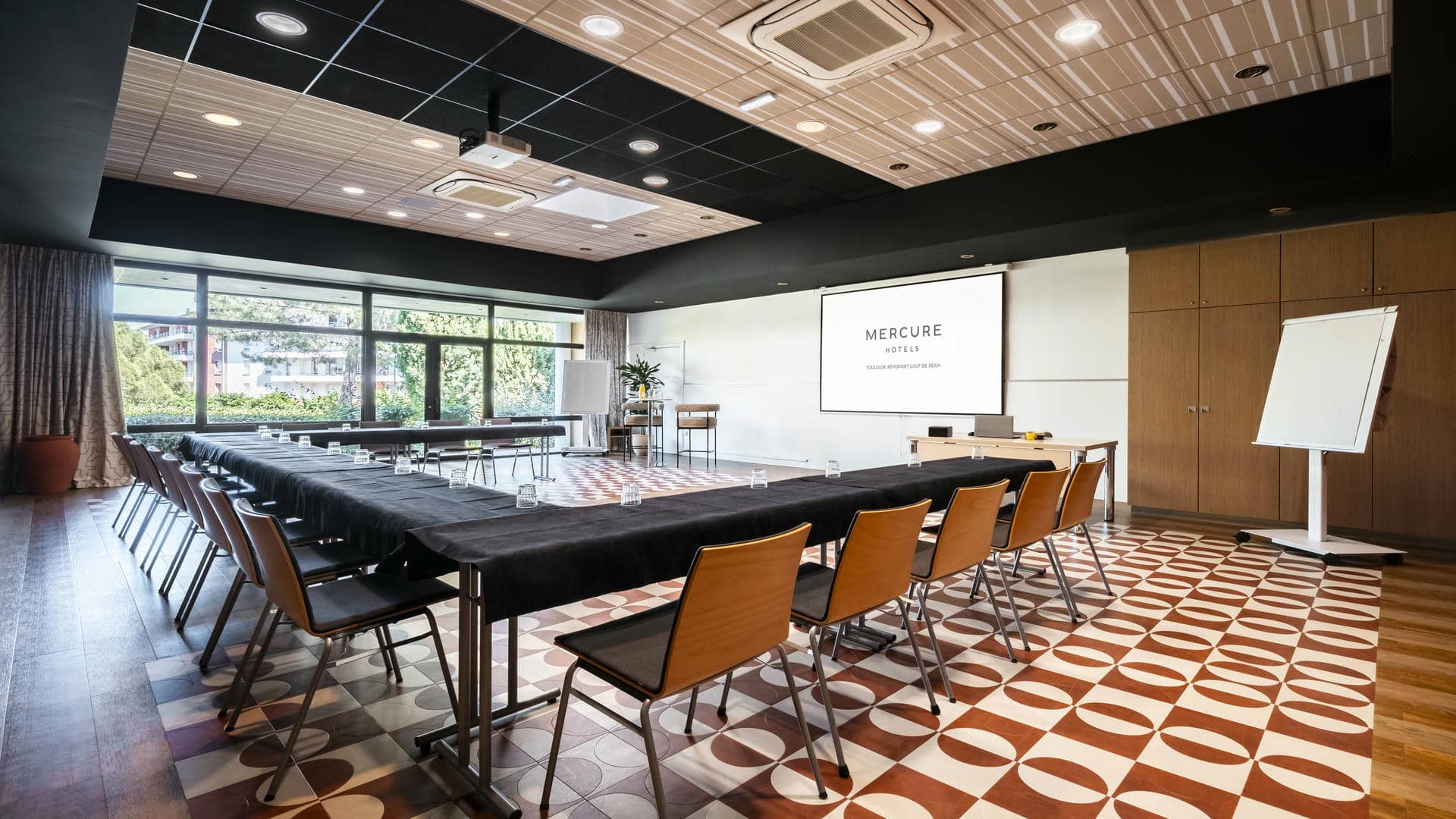 Salle de séminaire avec tables disposées en U et recouvertes de tissu noir, chaises hautes, mange-debout, écran mural, et vue sur la végétation et les bâtiments de l’hôtel séminaire.