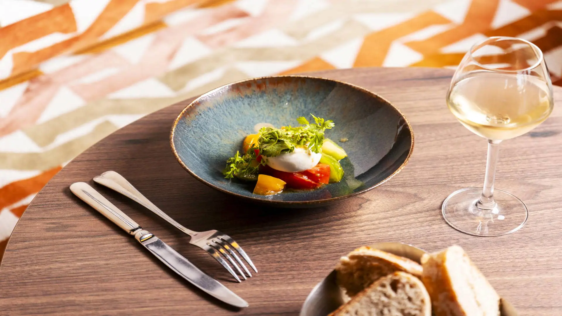 Plat A L’Epicerie Restaurant Experience de Toulouse Seilh, avec tomates anciennes, mozzarella artisanale et condiments, assise orangée, table en bois, verre de vin et corbeille à pain.