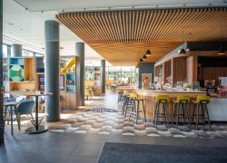 A droite, le bar dédié aux séminaires présente un comptoir et un plafond habillés de bois clair, accompagnés de hautes chaises jaunes. A gauche, la salle de restaurant est structurée par des étagères colorées faisant office de séparations, afin de créer des espaces plus intimes.