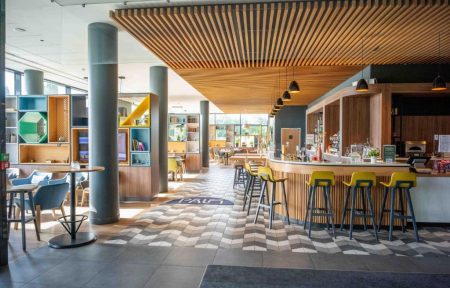 On the right, the seminar bar features a counter and ceiling clad in light wood, with tall yellow chairs. On the left, the restaurant area is divided by colourful shelving units, creating more intimate spaces.