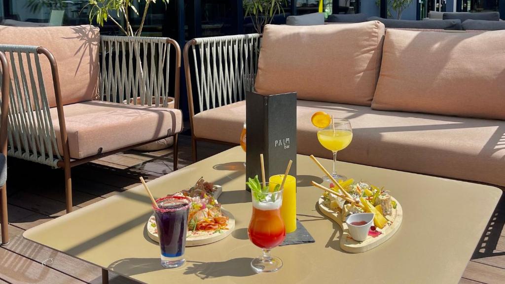 Metal-framed armchairs and sofas with stretched fabric are arranged on the hotel’s seminar terrace around a low table made of wooden planks. Appetizer boards, colourful cocktails and a PALM menu are placed on the table.