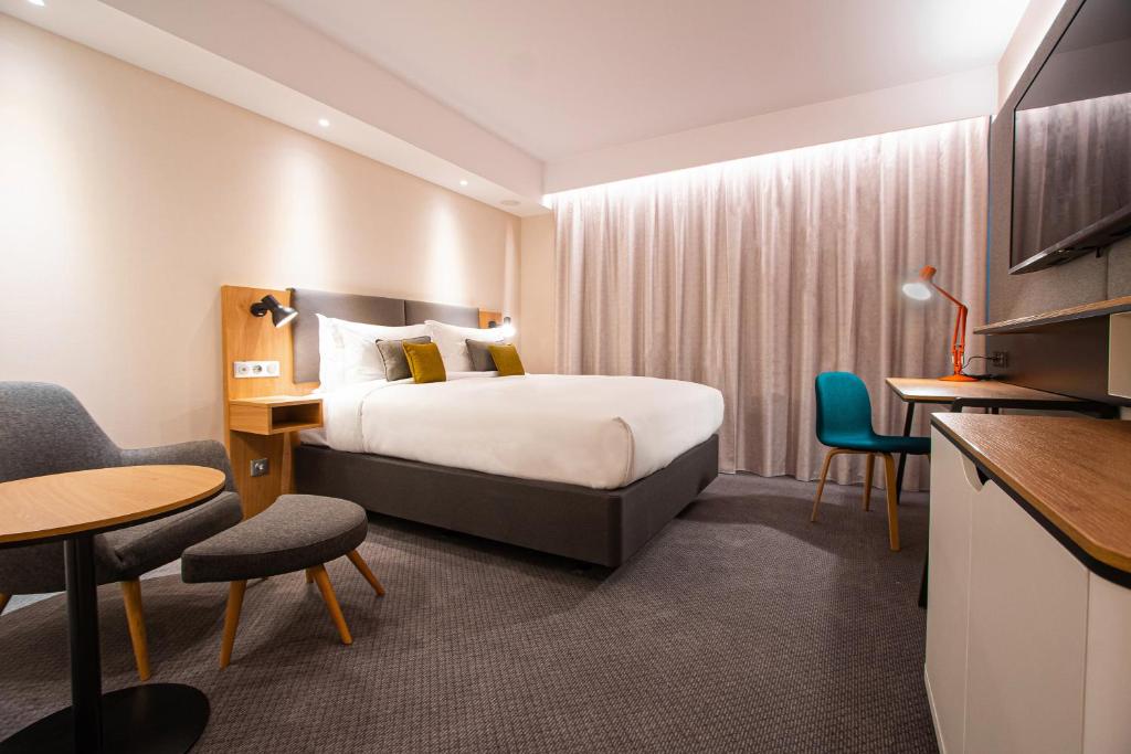 Bedroom with a double bed, two white pendant lights hanging at pillow height, thick beige curtains, a desk with a blue chair, a TV cabinet, a grey armchair with a footstool, and a round wooden table. A large Haussmann-style window overlooks a Parisian neighbourhood.
