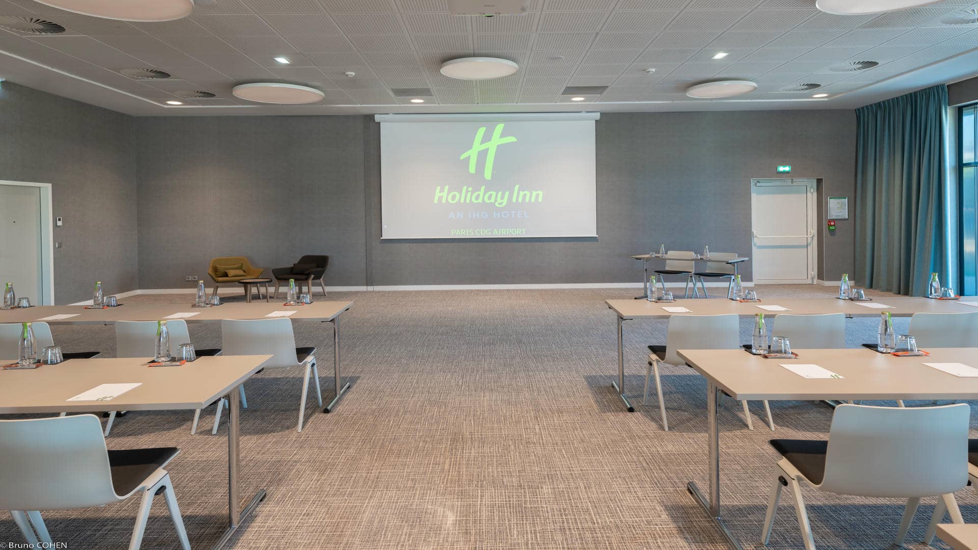 Seminar room with grey striped carpet, grey walls, green curtains, and a wall-mounted screen displaying “Holiday Inn Paris CDG Airport.” Desks are arranged classroom-style, with colourful armchairs placed beside the screen.