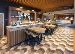 Le comptoir du bar, situé face à la réception de l’hôtel séminaire, est habillé de bois et bordé de tables en bois pour quatre personnes. Le sol associe carrelage marron et parquet.