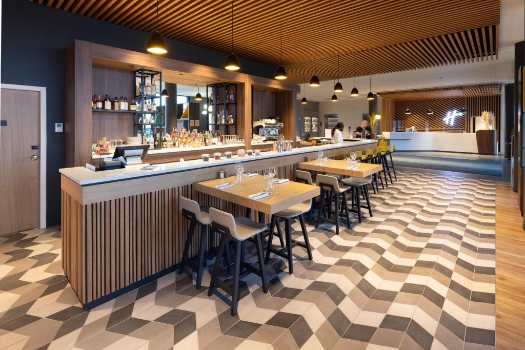 The bar counter, located opposite the seminar hotel reception, is clad in wood and lined with adjoining four-person wooden tables. Brown tiles and parquet flooring define the space.