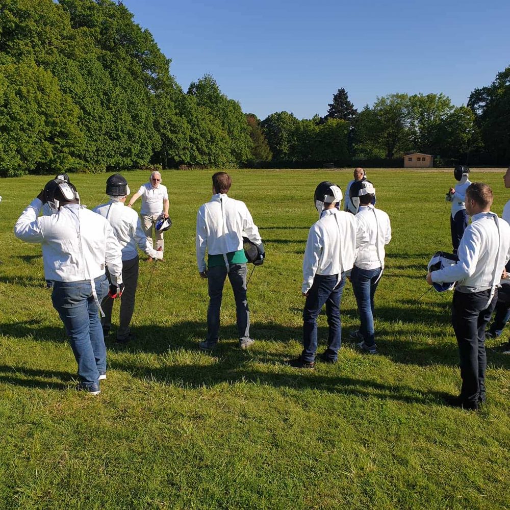 Activité extérieure adaptée pour un séminaire au vert, comprenant un groupe de personnes en vestes d'escrime blanches tenant masques et fleurets rassemblées sur une pelouse au premier plan, un instructeur en polo blanc au centre, et une ligne d'arbres verts denses sous un ciel bleu en arrière-plan.