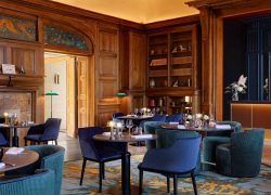 Restaurant avec plafond jaune en forme de dôme, murs habillés de boiseries avec comptoir intégré et fond bleu, parquet et tapis rond au sol, cheminée, lampes de bibliothèque vertes et livres sur les étagères.