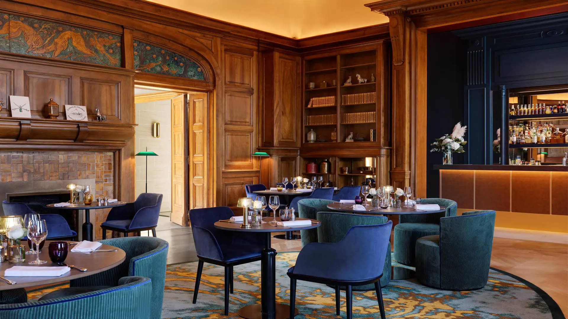 Restaurant with a yellow dome-shaped ceiling, wood-panelled walls with an integrated bar counter and blue back panel, parquet flooring, a round rug, fireplace, green library lamps and bookshelves.