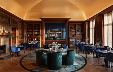 Bar with a yellow dome-shaped ceiling, wood-panelled walls with an integrated bar counter and blue back panel, parquet flooring, a round rug, fireplace, green library lamps and bookshelves.