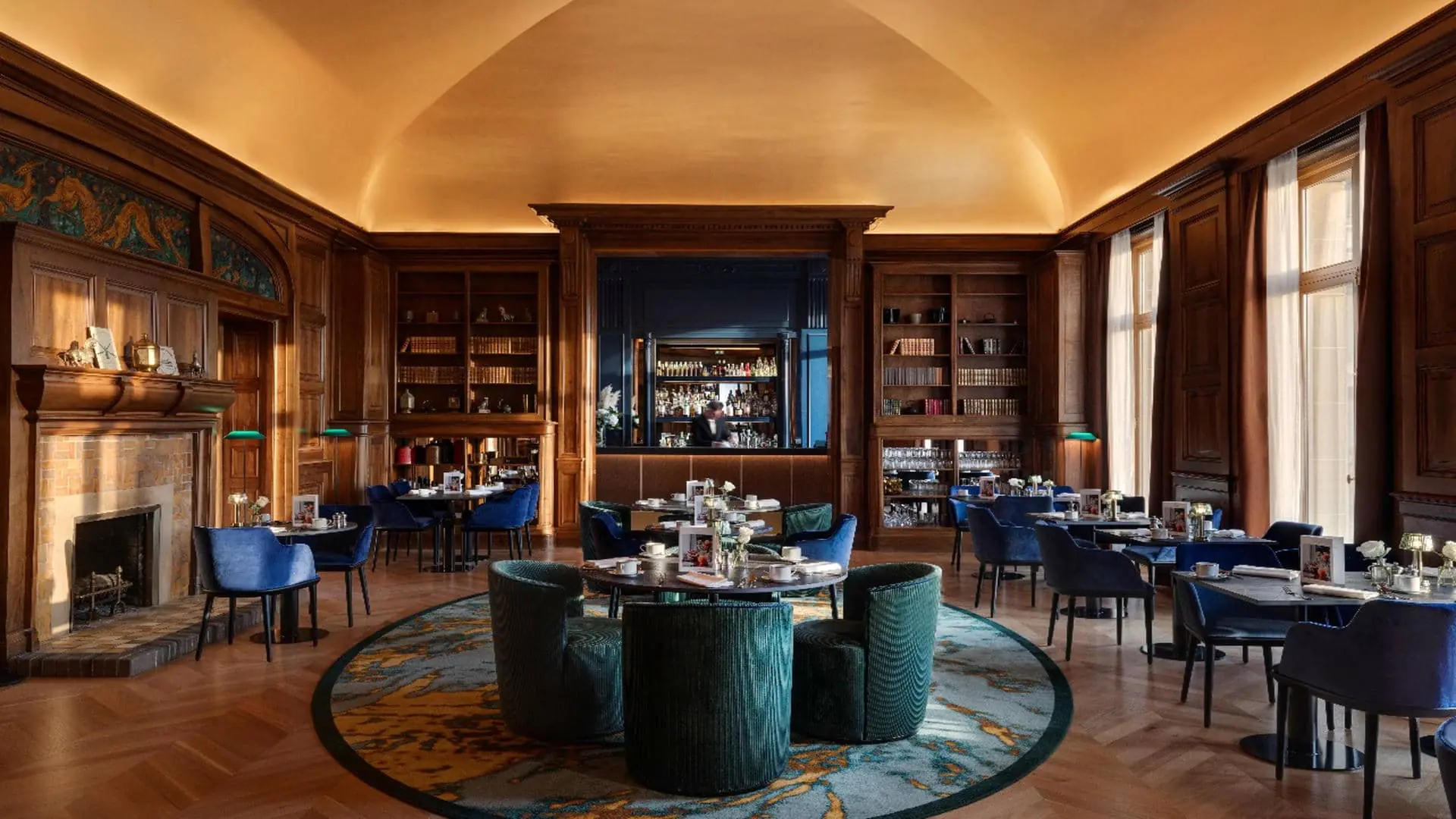 Bar with a yellow dome-shaped ceiling, wood-panelled walls with an integrated bar counter and blue back panel, parquet flooring, a round rug, fireplace, green library lamps and bookshelves.