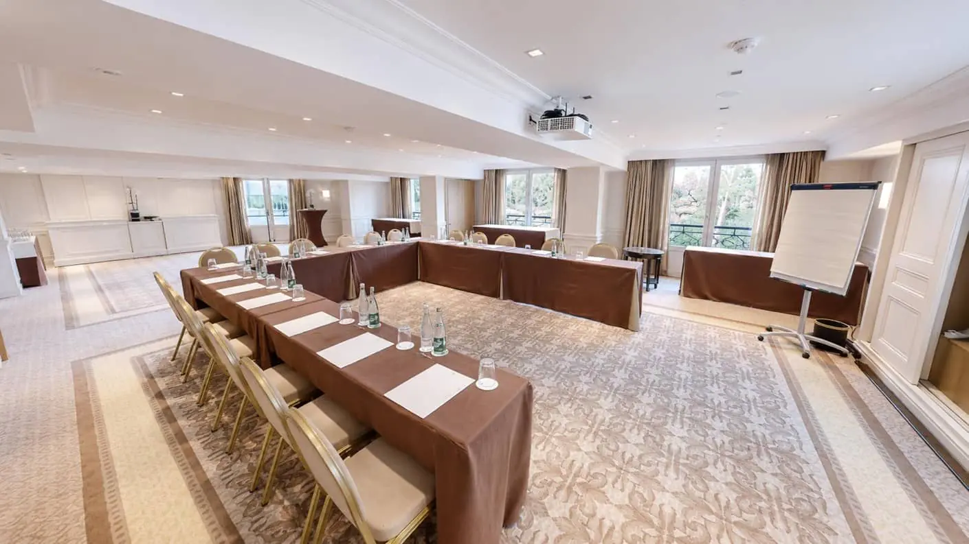 Seminar room rental with royal-patterned brown carpet, cream and gold chairs, U-shaped meeting tables with brown tablecloths, four French doors and a white break station at the back of the room.