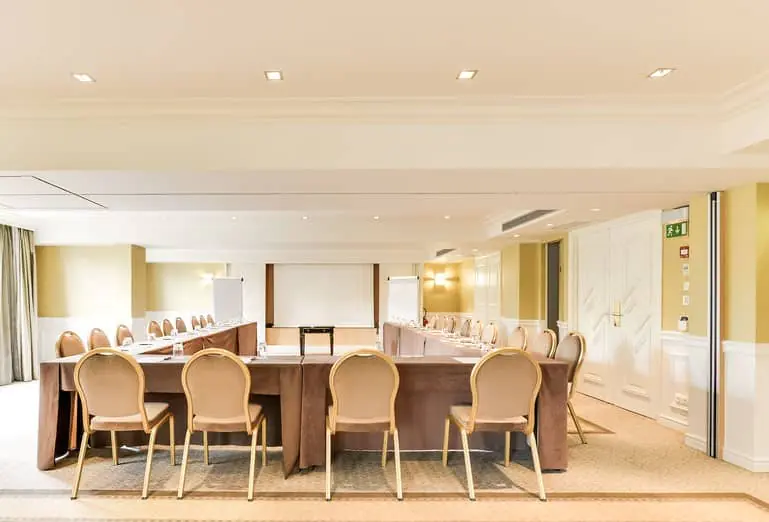 Meeting room with U-shaped tables, light carpet flooring, white and green walls, white ceiling and a projection screen.
