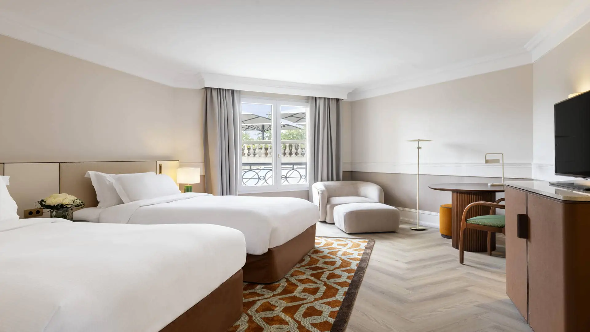 Twin beds on a rug with ring patterns, facing a TV unit, with an armchair and footrest, table and chair, and pouf, in a very bright room overlooking the upper terrace.