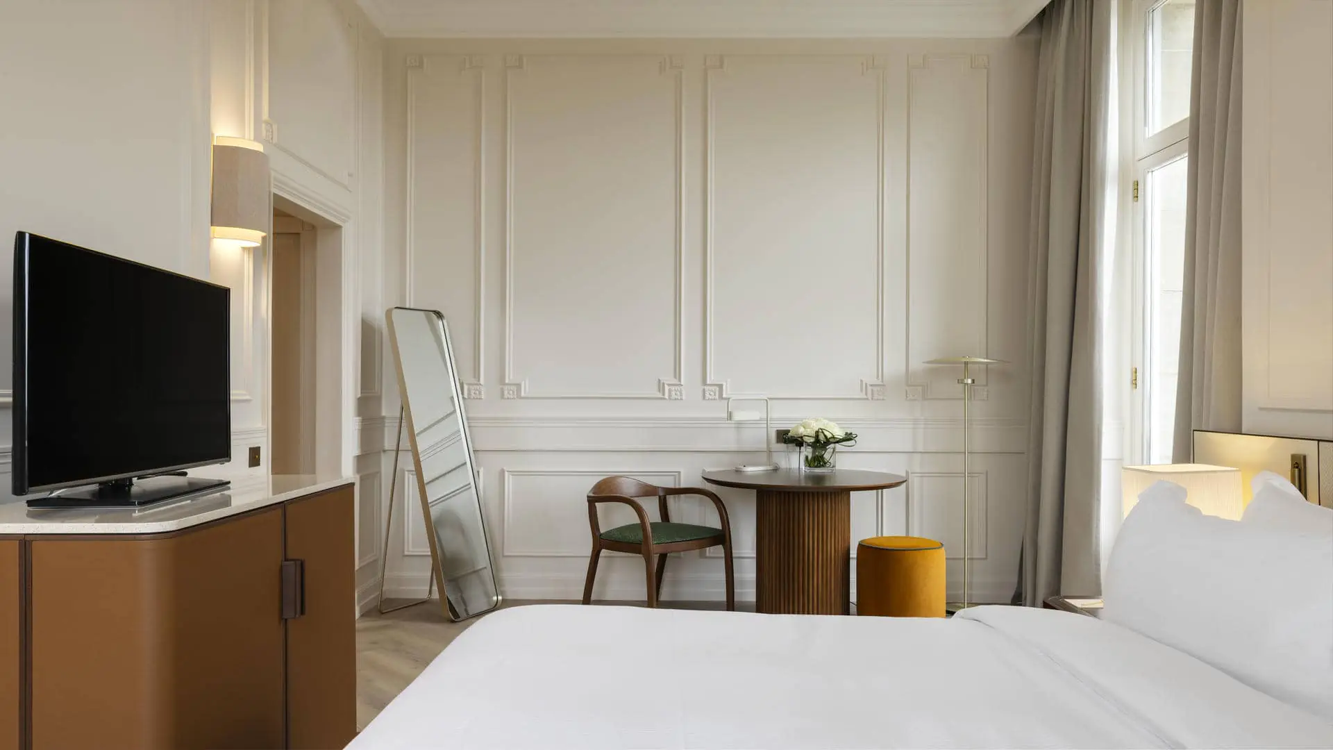 Twin beds on a rug with ring patterns, facing a TV unit, with an armchair and footrest, table and chair, and pouf, in a very bright room overlooking the upper terrace.