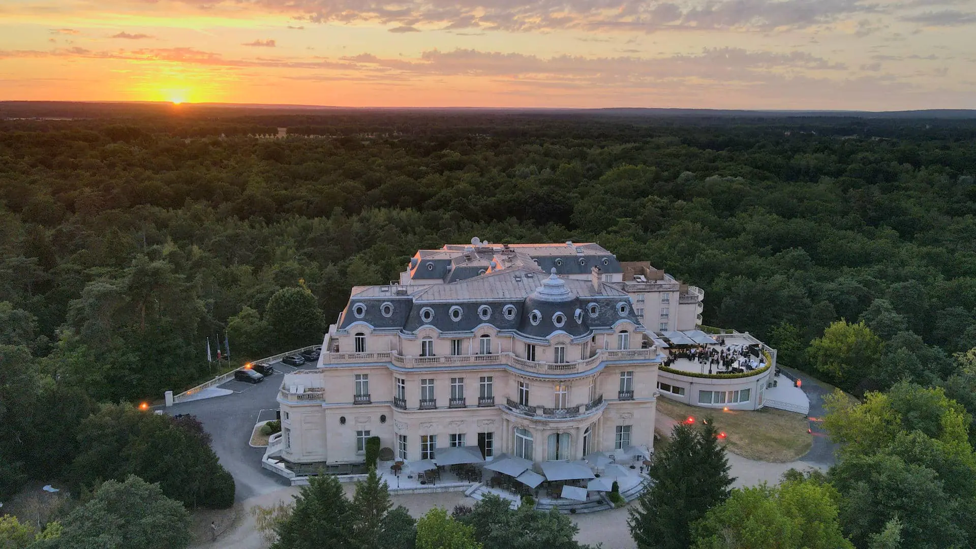 Château façade surrounded by the Chantilly forest at sunset, with views of the ground-floor terrace, upper terrace and parking area behind the buildings.