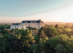 Façade du château entourée par la forêt de Chantilly, ciel rosé et brume sur la canopée.