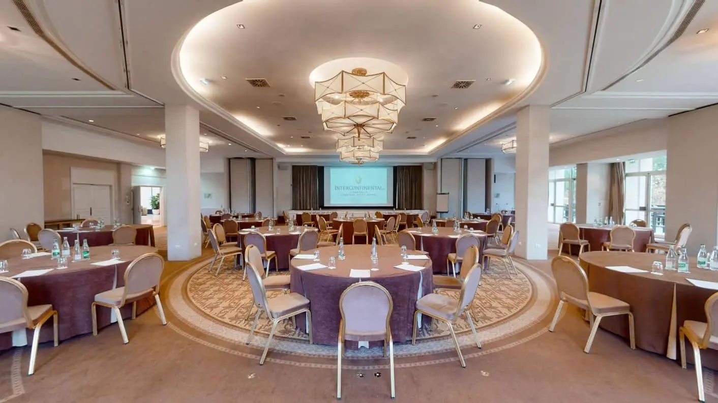 Convention room with a stage, beige carpet, round tables, a large projection screen and chandeliers on the ceiling.