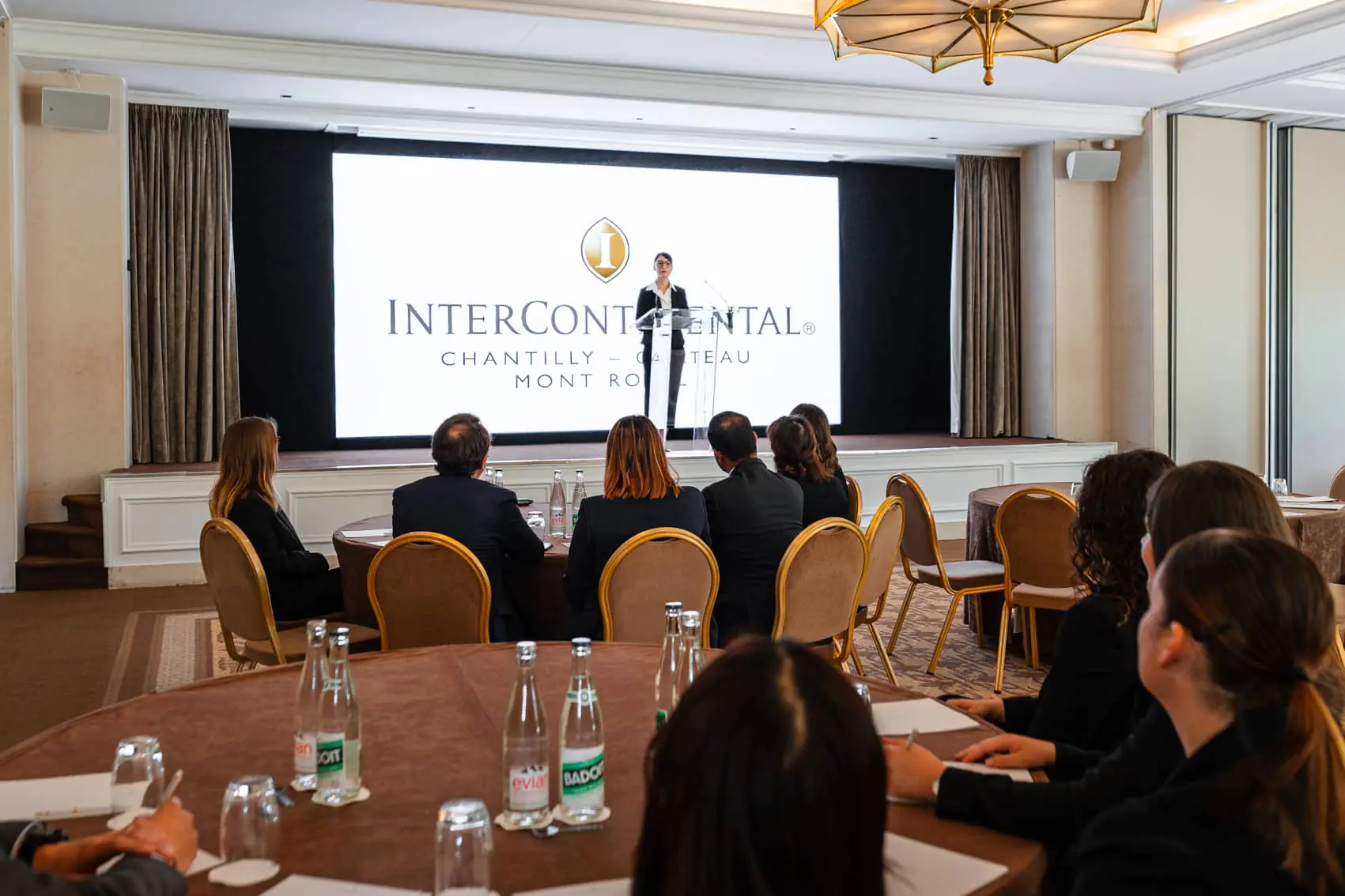Convention room with a stage, beige carpet, round tables and a large projection screen.
